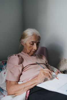 Elderly woman in pink sweater writing on paper while sitting on bed in a cozy room.