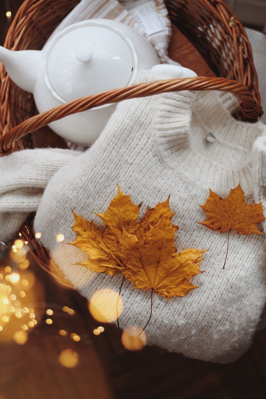 Warm autumn scene with a knitted sweater, maple leaves, and a tea pot in a woven basket.