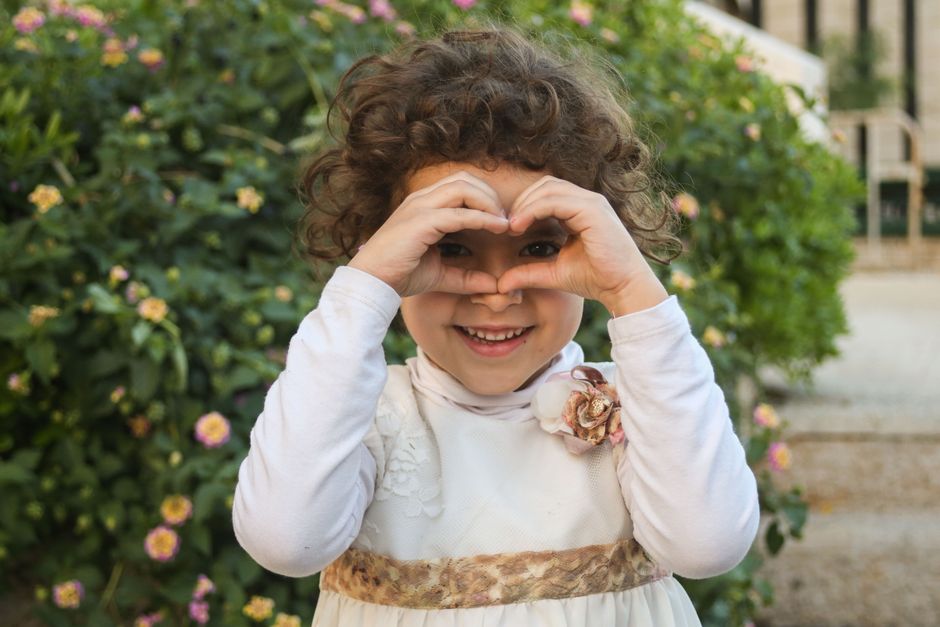 A happy curly-haired girl in a white dress smiling and making a heart shape with her hands in a garden