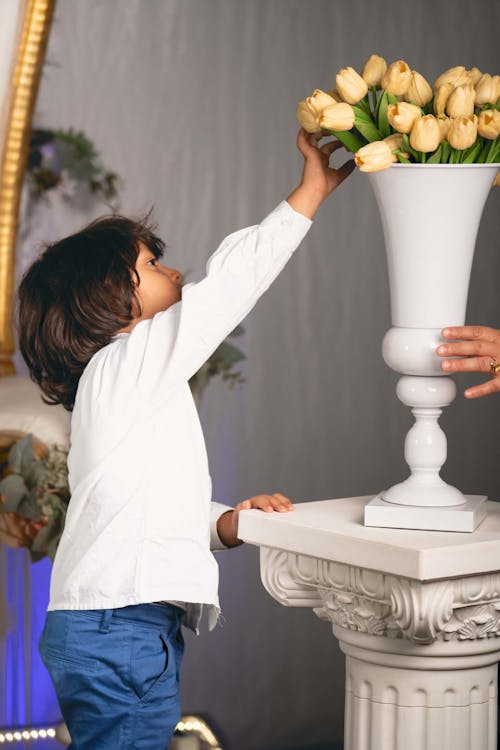 A Young Boy in White Long Sleeve Shirt Touching a Yellow Tulip Flowers on White Vase