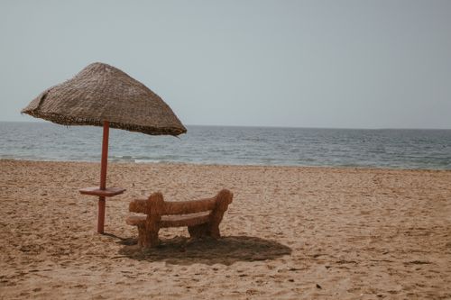 Free Quiet beach scene featuring a rustic wooden bench and straw umbrella by the seaside. Stock Photo