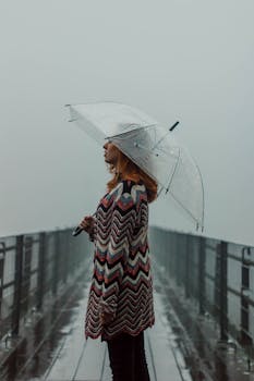 Woman standing on a foggy pier holding a clear umbrella, creating a moody and serene scene.