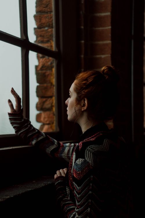Free A woman in a patterned sweater gazes thoughtfully out a large window. Stock Photo