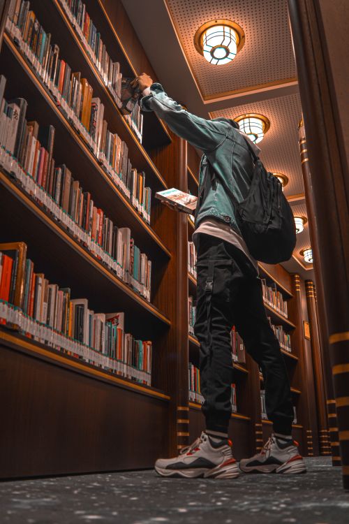 Free A man with a backpack reaches for a book in a classic library interior. Stock Photo