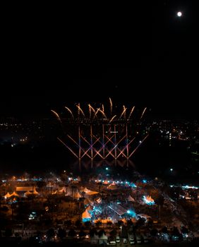 Aerial view of vibrant fireworks illuminating a bustling night festival below the city sky.