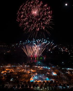 A vibrant fireworks display illuminates the night sky above an urban festival, creating a festive atmosphere.