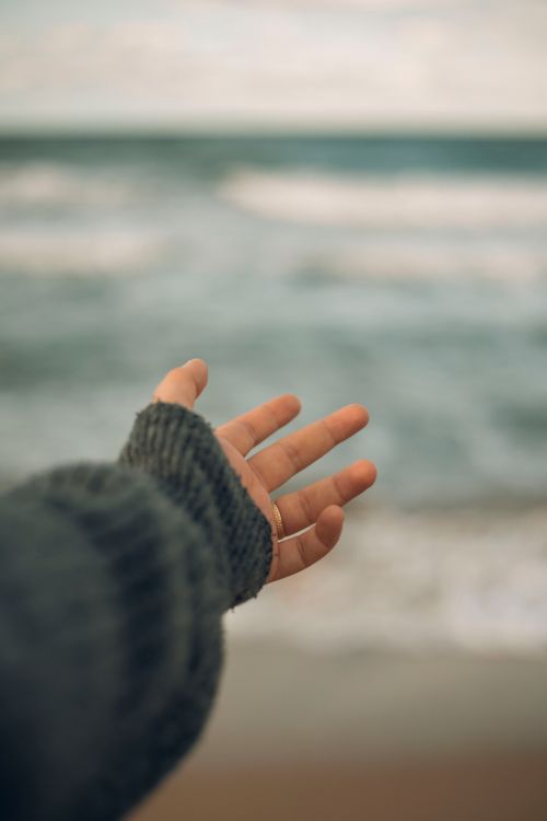 Free A hand in a sweater reaching out towards the ocean on a cloudy day. Stock Photo