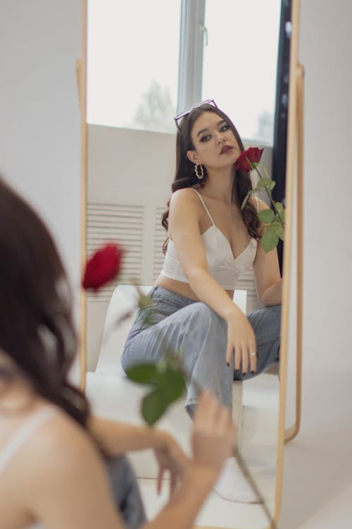 A Woman in White Spaghetti Strap Top Sitting in Front of the Mirror while Holding a Rose