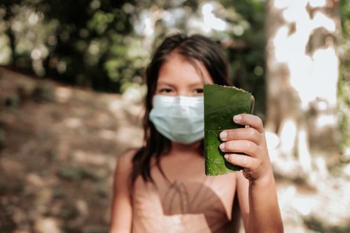 Free A young girl wearing a face mask holds a green leaf in a forest setting, reflecting nature and new health norms. Stock Photo