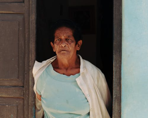 An Elderly Man in Blue Shirt Standing Near Brown Wooden Door