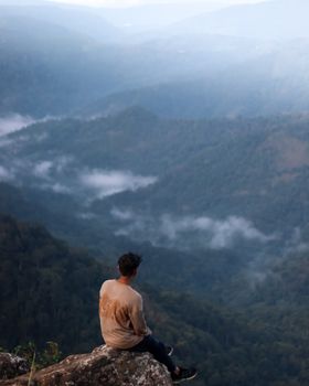 A man sits on a cliff in Vagamon, India, enjoying a misty mountain view, showcasing adventure and tranquility.