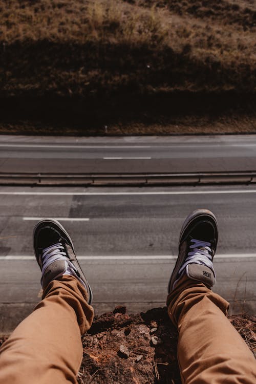 A Person Wearing Black Sneakers Sitting on a Cliff with the View of an Empty Road