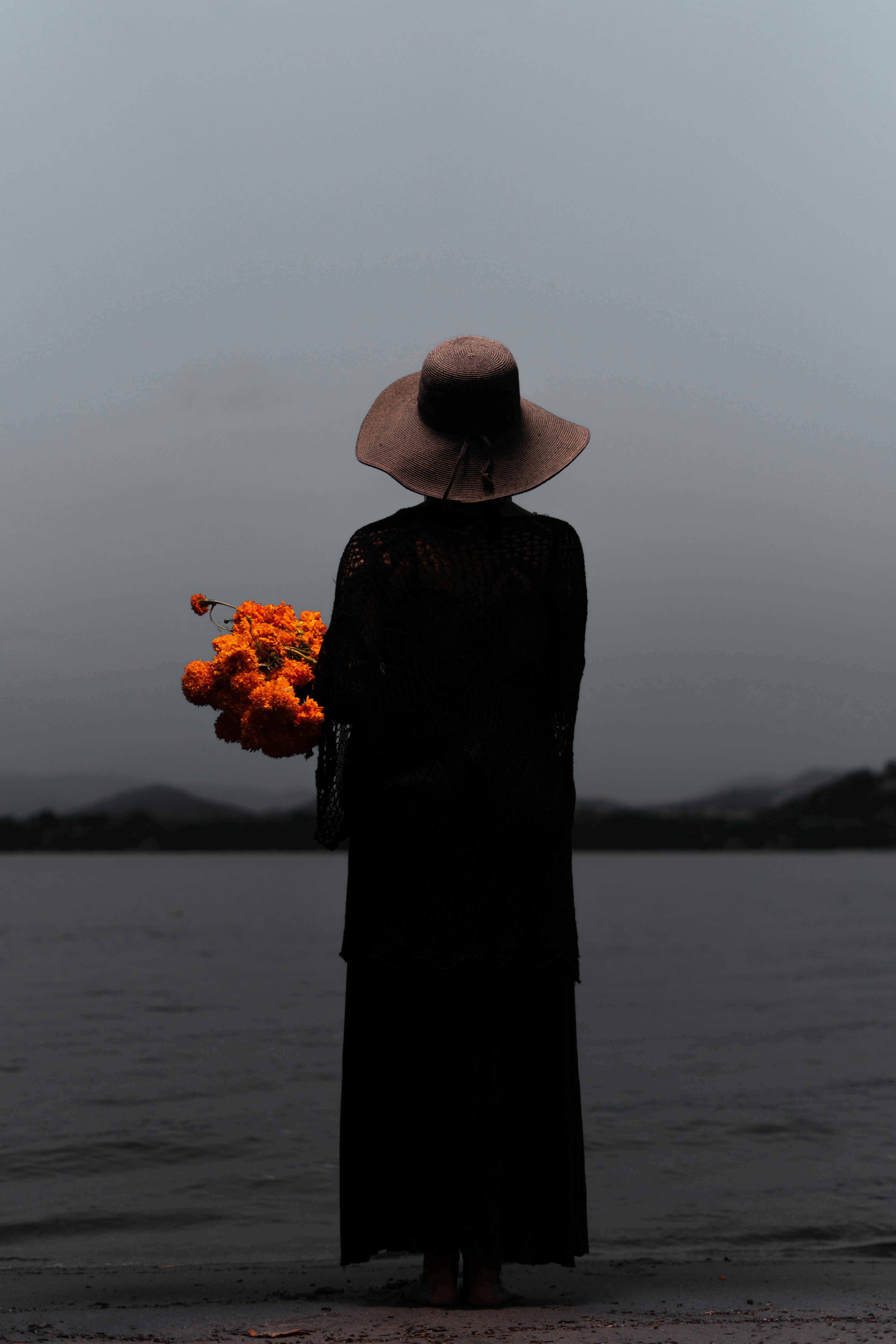 A person in a black dress holds marigolds near the shore in Acapulco at twilight.