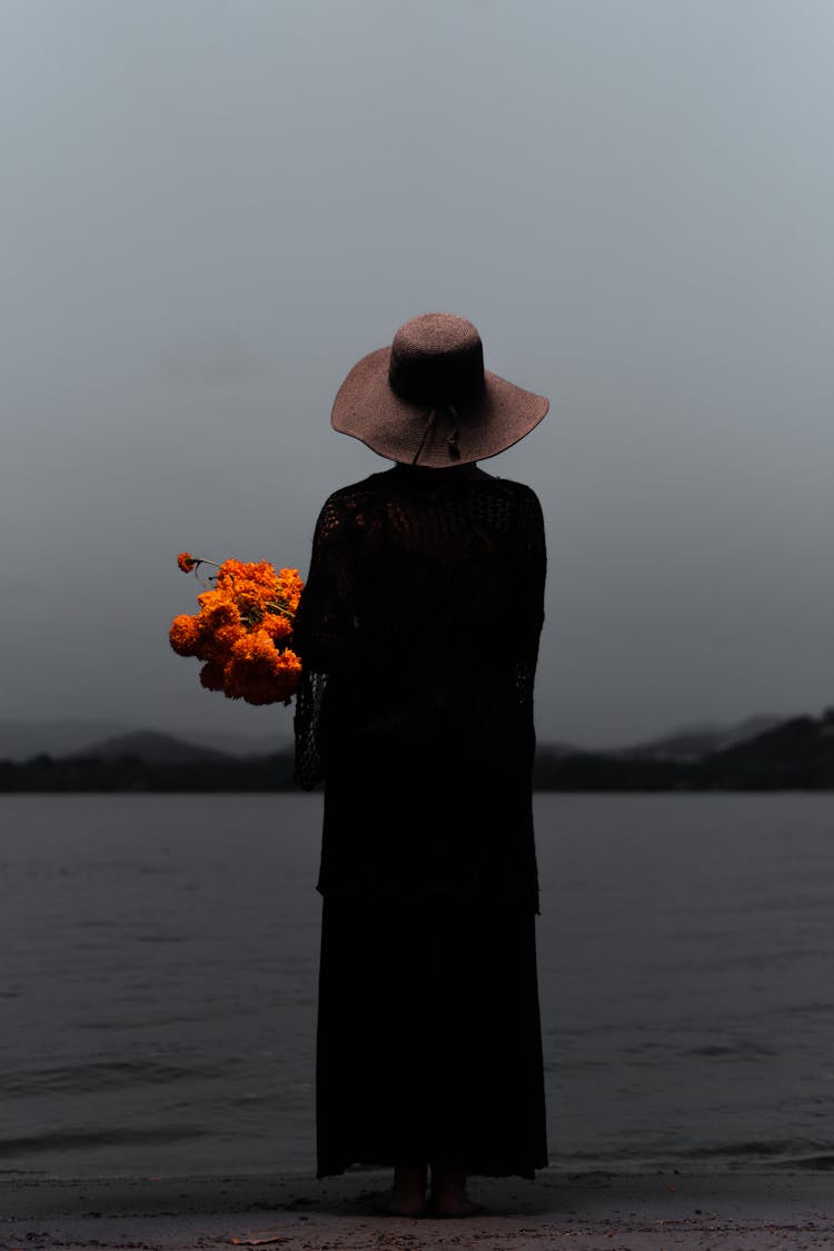 Back View Shot Of Person In Long Black Dress Holding A Flower While Standing On The Shore Of A Beach