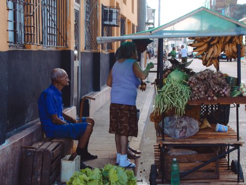Free A vibrant street market stall selling fresh produce with two adults interacting on a sunny day. Stock Photo