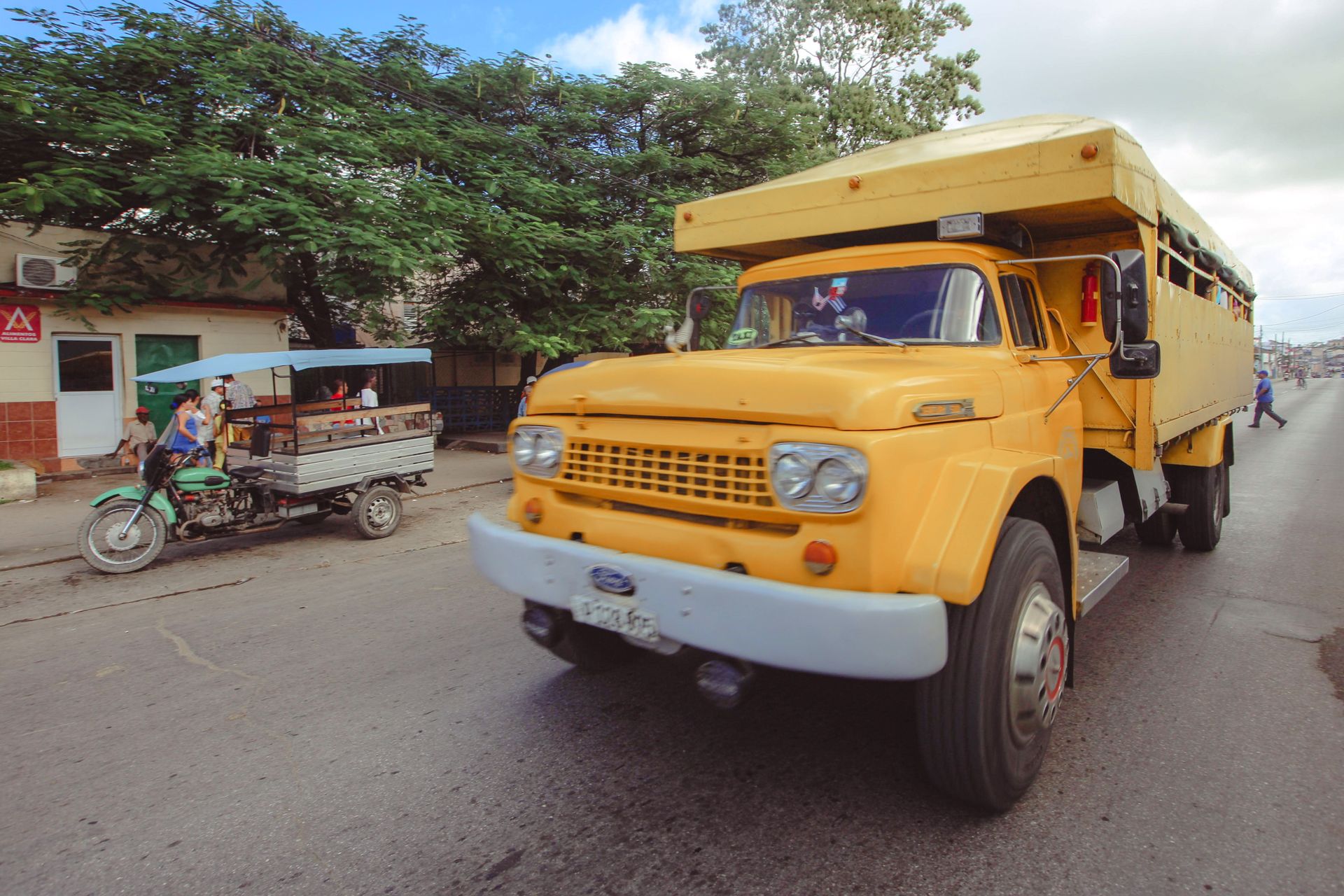 A vibrant yellow truck driving on an urban street with a motorcycle taxi nearby.