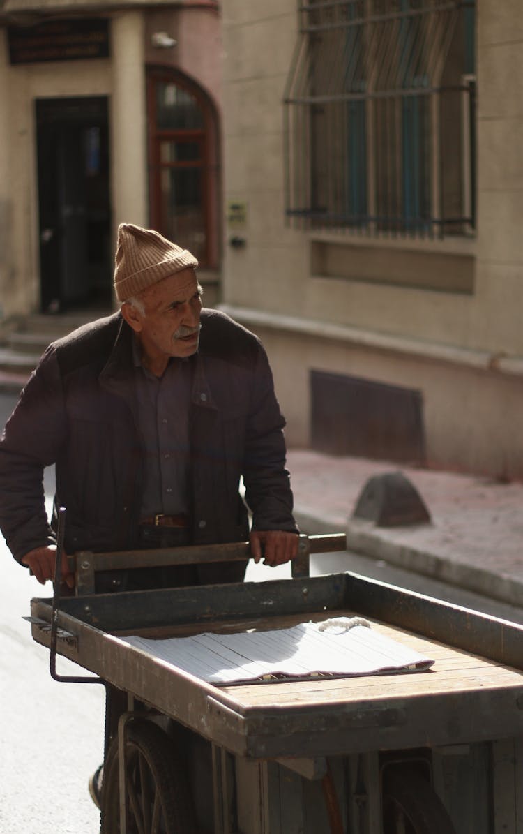 Senior Man Walking In Brown Coat And Beanie Pushing A Wooden Trolley Cart On Street