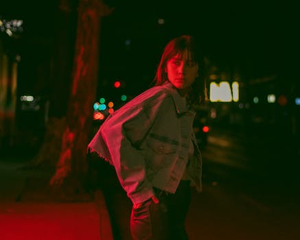 A young woman in a denim jacket stands on a dimly lit street at night, Ciudad de México.