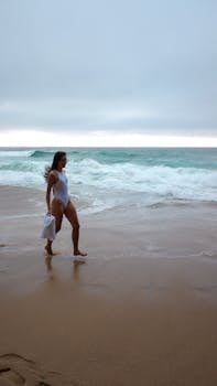 A woman strolls along a serene beach in a white swimsuit, capturing coastal tranquility.