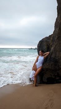 Woman in white swimsuit relaxing on rocky beach against stunning ocean backdrop.