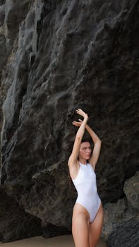 A woman in a white swimsuit poses gracefully against a rugged rock formation outdoors.