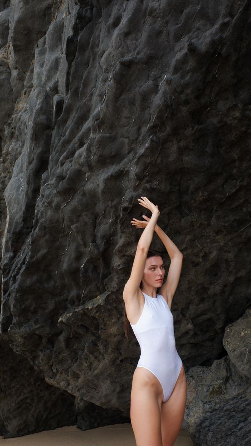 Free A woman in a white swimsuit poses gracefully against a rugged rock formation outdoors. Stock Photo