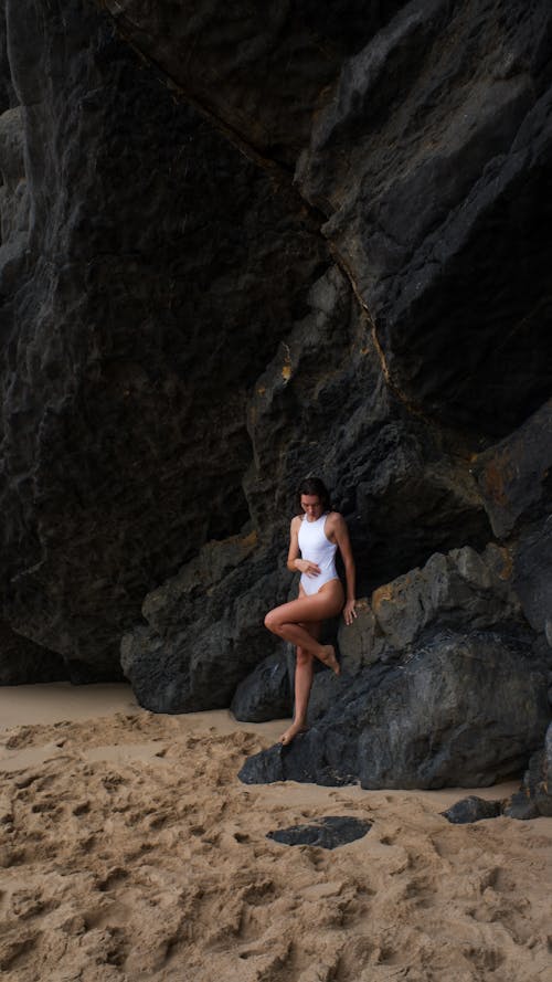 A Woman in White Swimsuit Sitting on the Rock Near the Beach Sand