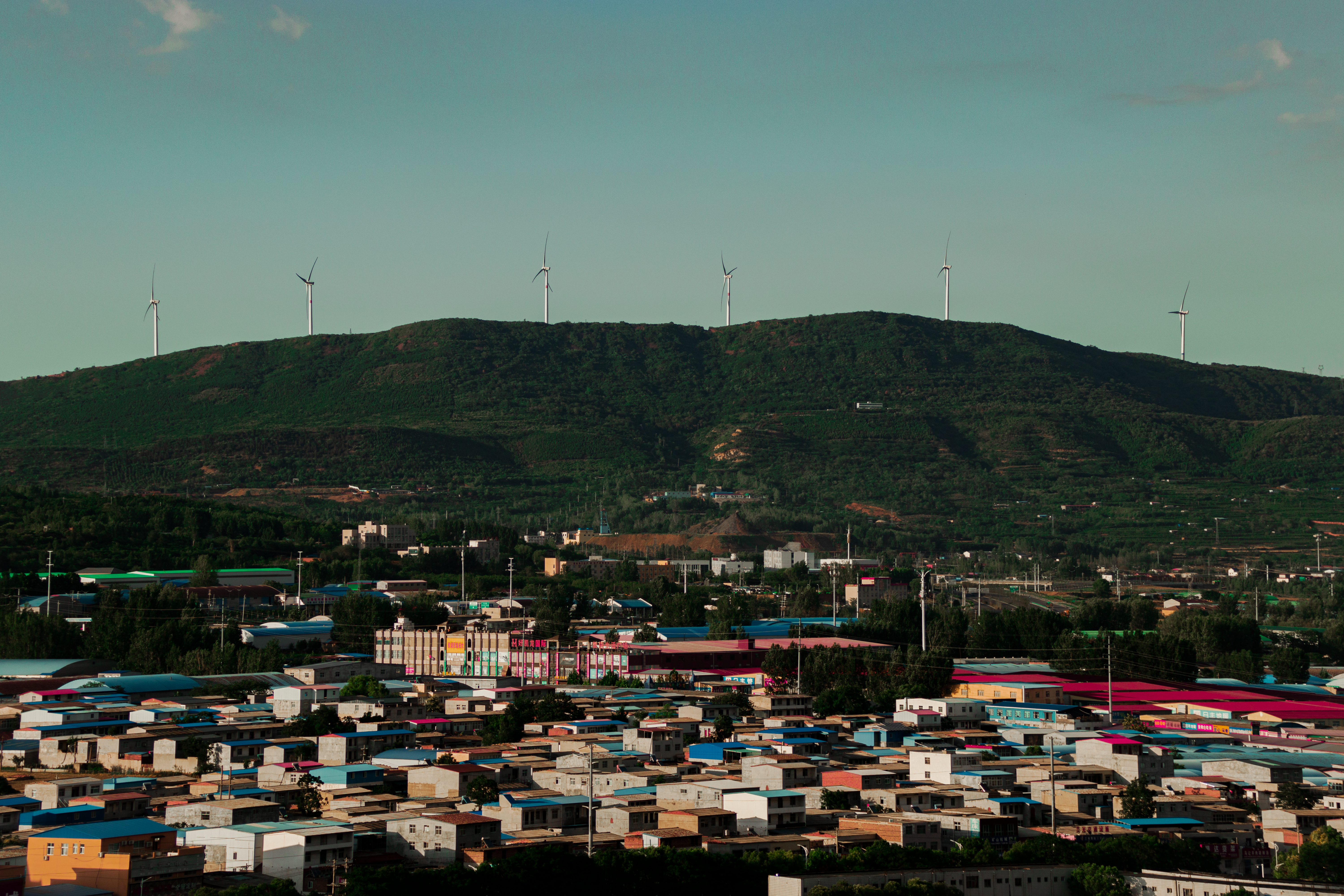 City view with wind turbines on a green hill, showcasing renewable energy at sunset.