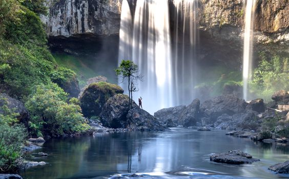A lone hiker standing on rocks near a mesmerizing waterfall surrounded by lush greenery.