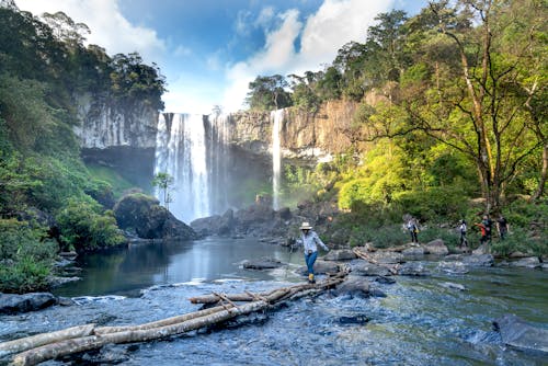 Woman Crossing the River on Brown Log Near Waterfalls