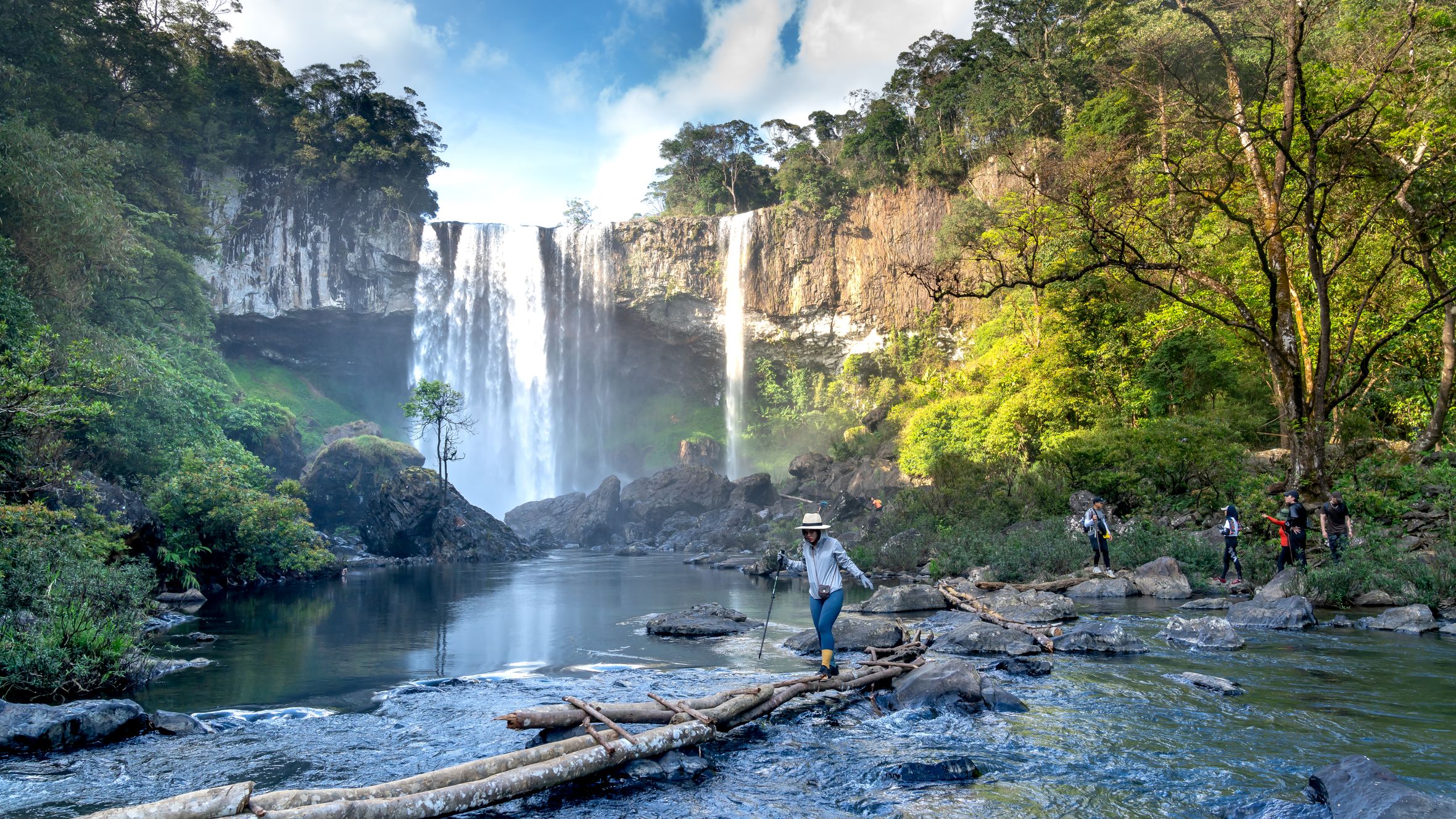 Group of hikers crossing a river with a stunning waterfall backdrop, surrounded by lush forest.