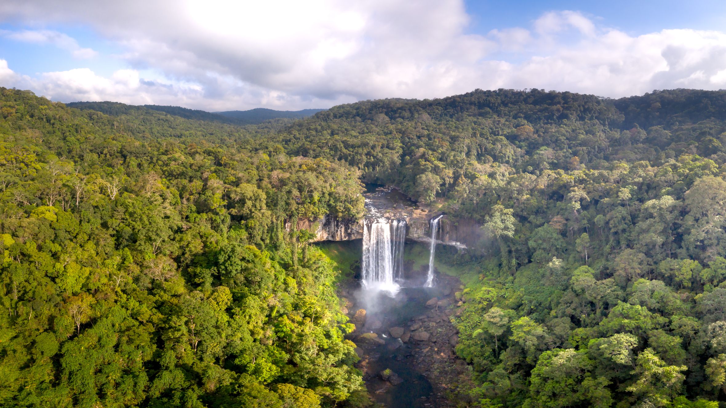 Stunning aerial view of a waterfall cascading through lush forest under a blue sky.