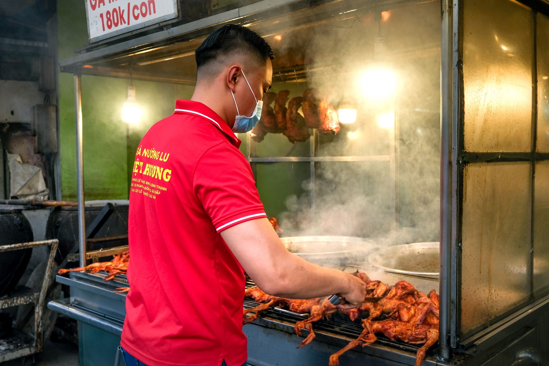 Street vendor grilling barbecue chicken on a busy street, wearing a face mask for safety.