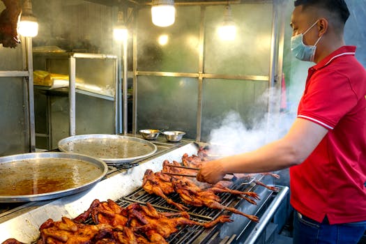 A man in a mask grills chicken in a smoky street food stall wearing a red polo shirt.
