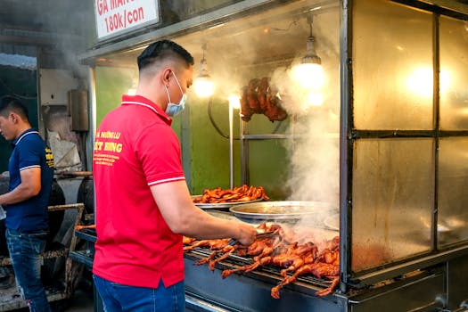 Asian man grilling chicken at a street food stall with smoke and face mask.
