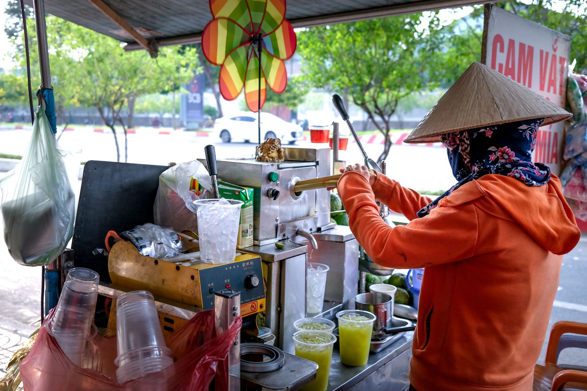 Street vendor wearing conical hat preparing fresh juice at outdoor cart with various ingredients.