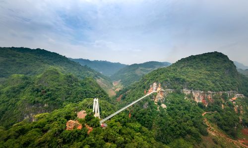 Free A panoramic view of a suspension bridge spanning lush green mountains under a cloudy sky. Stock Photo
