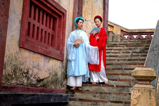 Two women in traditional Vietnamese dresses holding hand fans on ancient stairs.