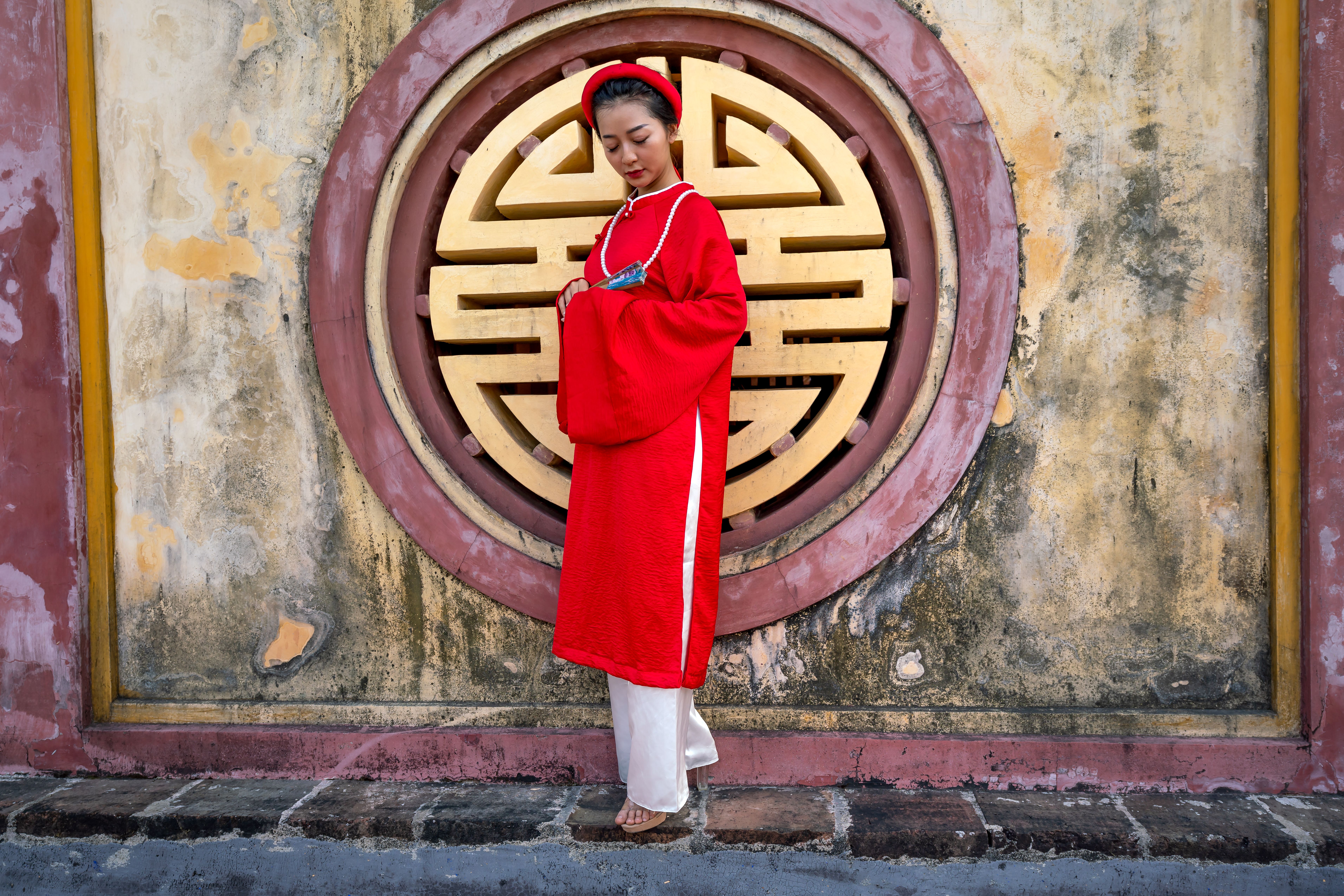 An Asian woman in a red ao dai stands gracefully against a vintage wall with a circular design.