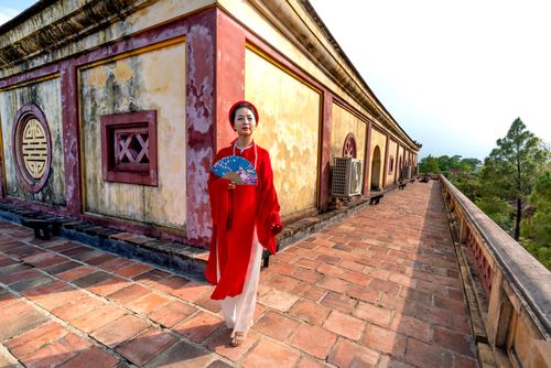 Free A woman in a traditional red ao dai stands in front of a historic building, symbolizing Vietnamese culture. Stock Photo