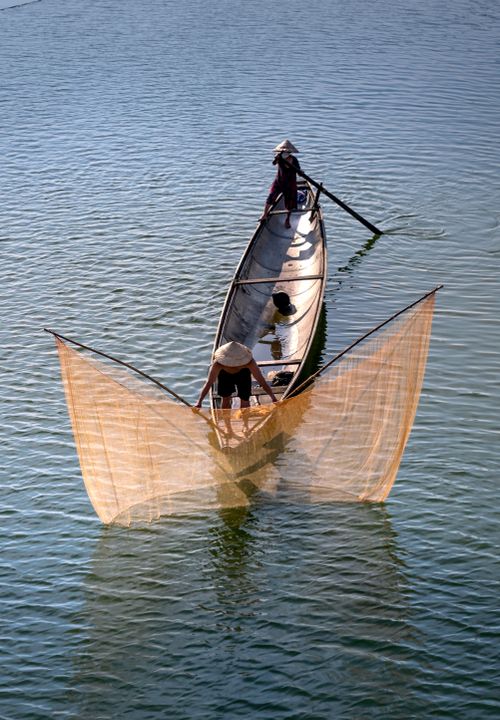 Free Aerial shot of fishermen in conical hats casting nets from a boat on serene blue waters. Stock Photo
