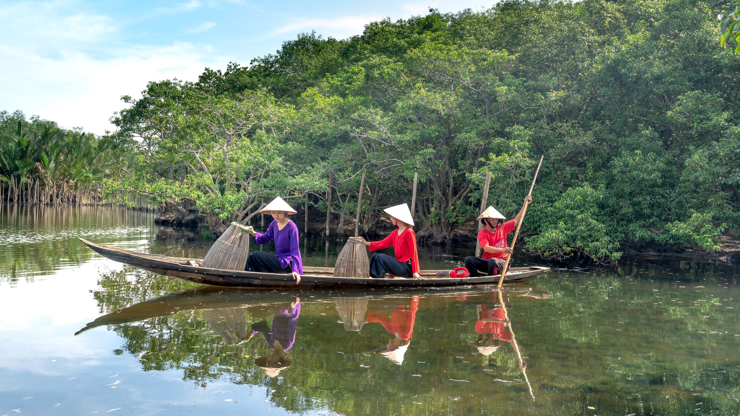 Asian fishermen navigate a river in a wooden boat, surrounded by lush mangroves.