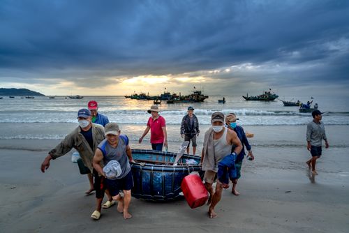 Free Group of fishermen carrying a haul on a beach during sunset with boats in the background. Stock Photo