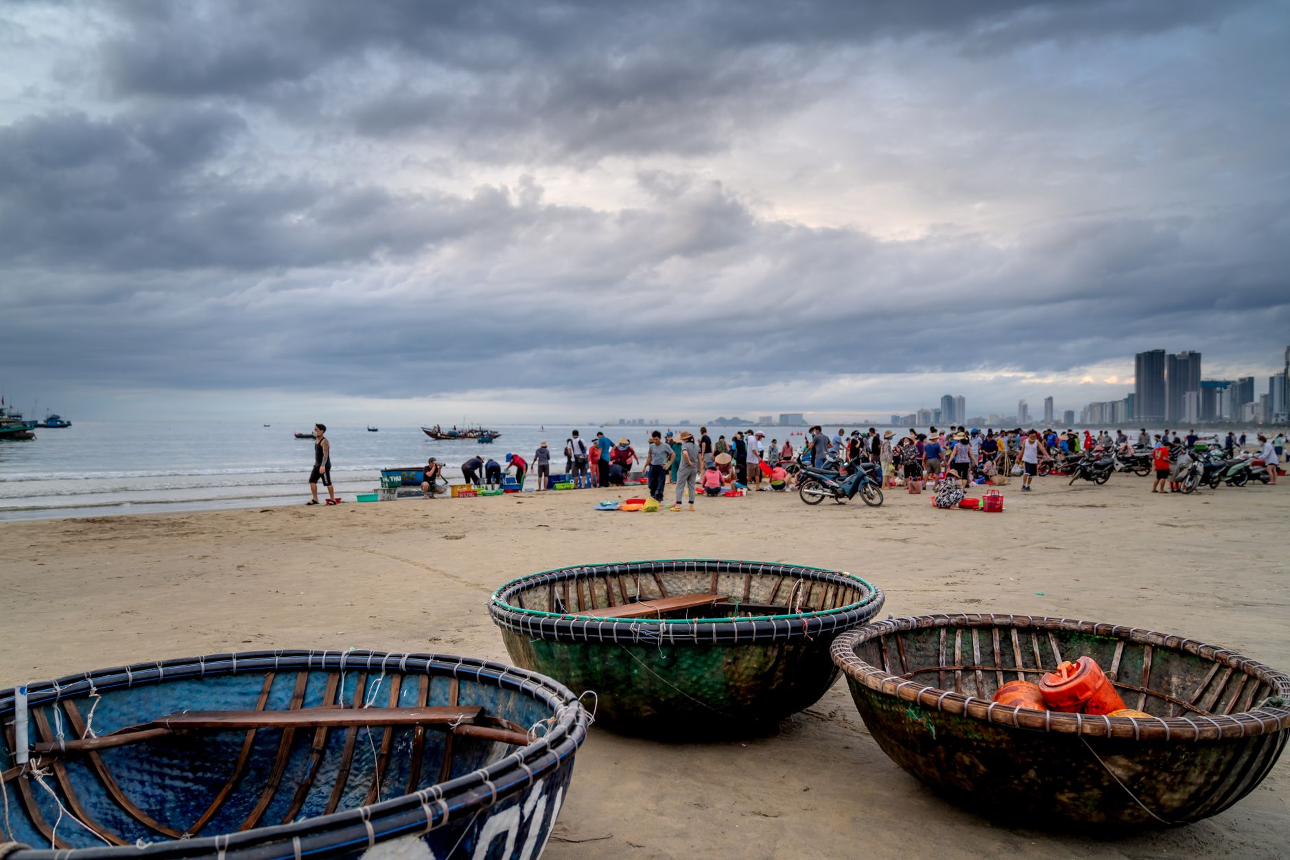 A bustling Vietnamese beach scene with traditional basket boats, people, and a cloudy sky.