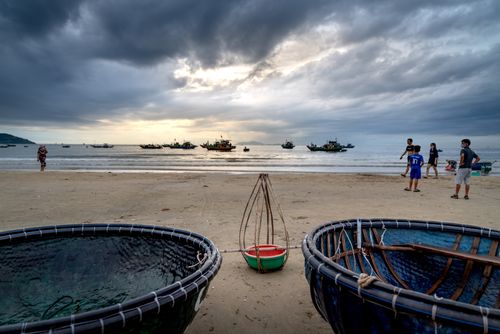 Free Beach scene with fishing boats near shore, under dramatic cloudy skies and people walking. Stock Photo