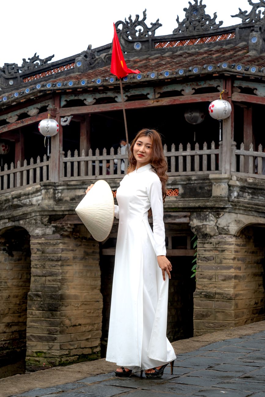 A woman in a white dress and traditional hat poses by a historic Japanese Bridge.