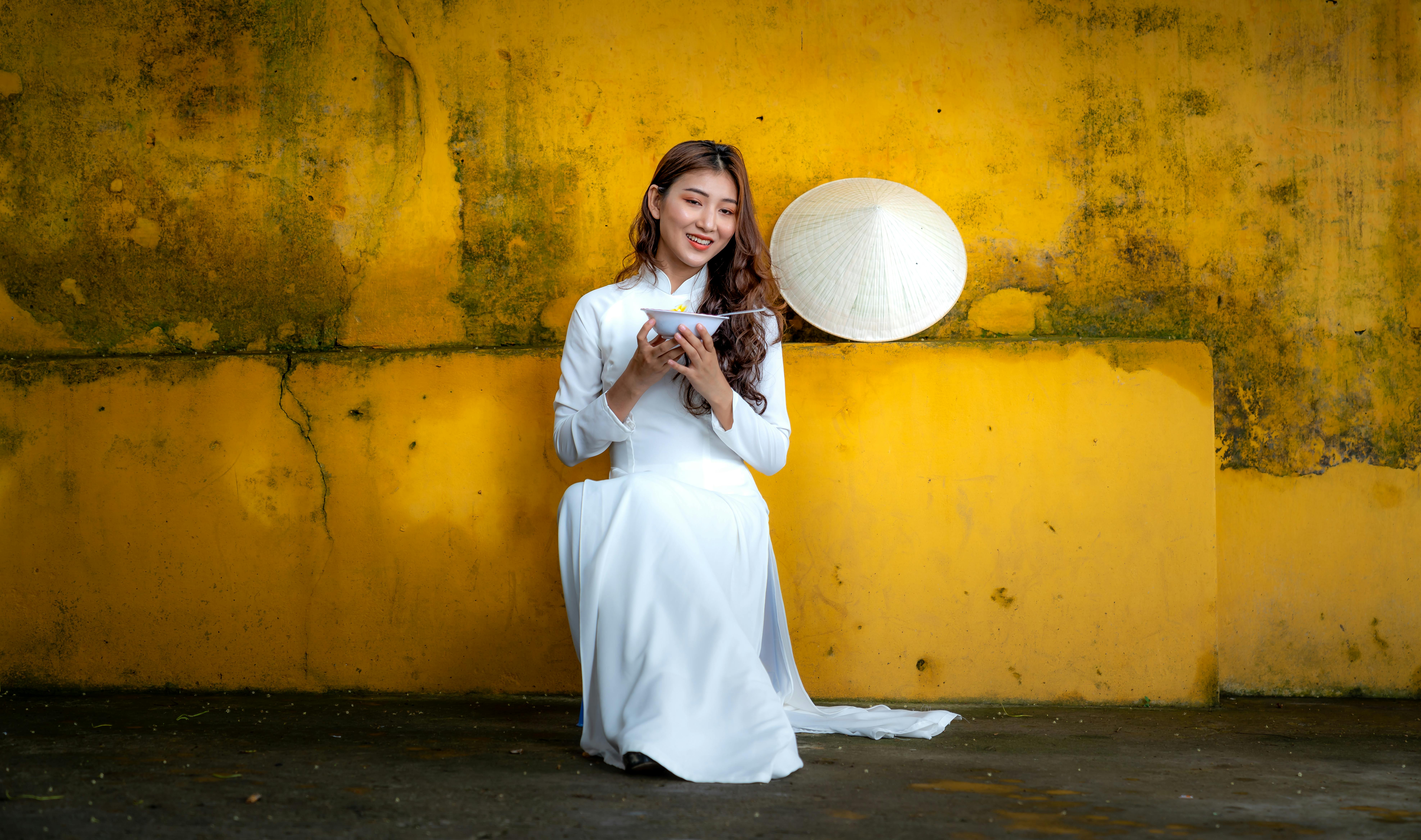 Portrait of an Asian woman in traditional dress smiling while holding a bowl, set against a vibrant yellow wall.