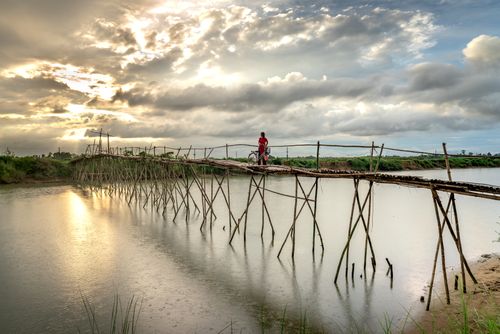 Free A person pushes a bicycle across a wooden bridge over a calm lake under a dramatic cloudy sky. Stock Photo