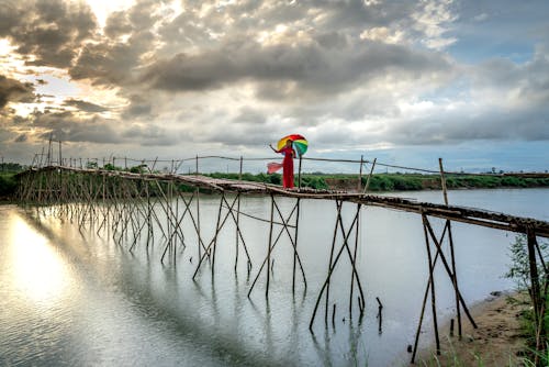 A Woman in Red Dress Standing on a Wooden Bridge while Holding an Umbrella