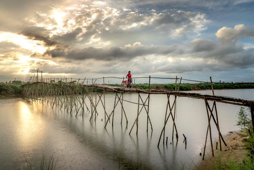 Free A woman pushes her bicycle across a rustic wooden bridge over a serene river at sunset. Stock Photo